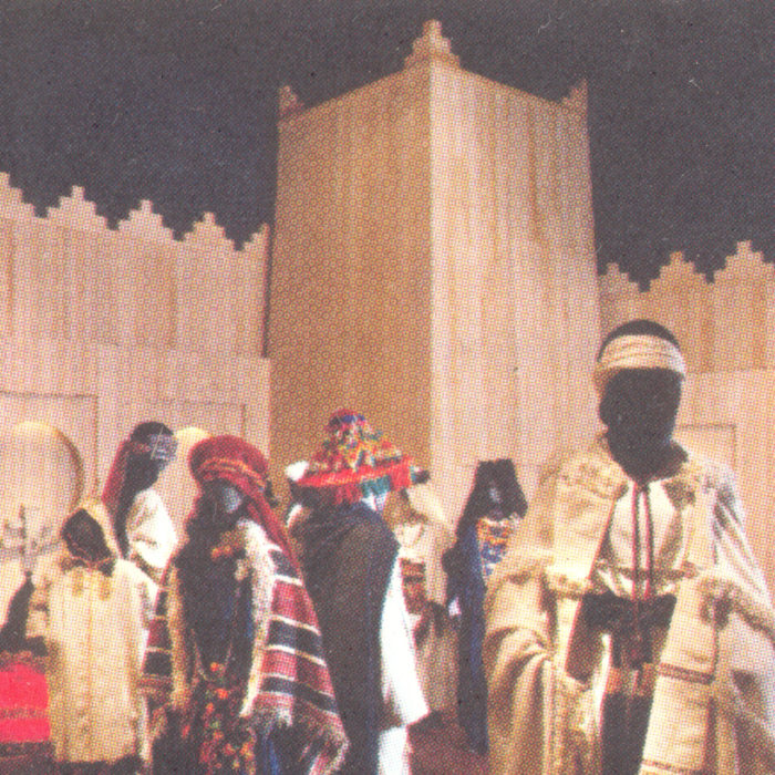 Scan of an image of a group of figures in traditional Berber dress, beige and gold fabric with colourful patterns sat against a wooden architectural structure of Berber provenance.