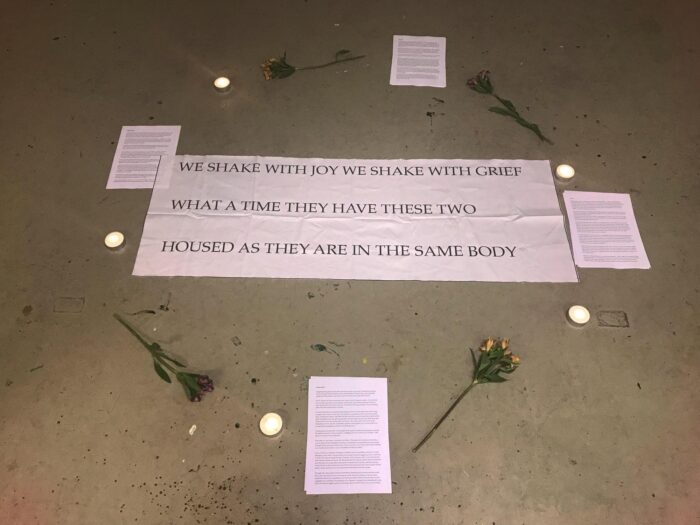 Photo of a grey/brown concrete floor with 4 separate piles of A4 sheets alongside single flowers and tea light candles in a circle that surround A4 sheets of white paper pasted together that read “we shake with joy, we shake with grief, what a time they have these two, housed as they are in the same body”