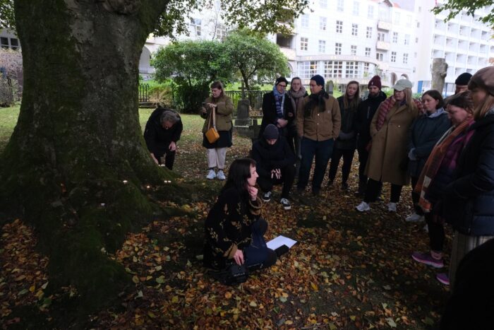 Photo of a large dark mossy tree in the foreground to the left in front of white buildings in the background, to the right of the tree is a figure sitting on the floor surrounded by orange autumnal leaves and a group of 13 people standing and listening. Two people bend down to put tea lights at the foot of the tree.